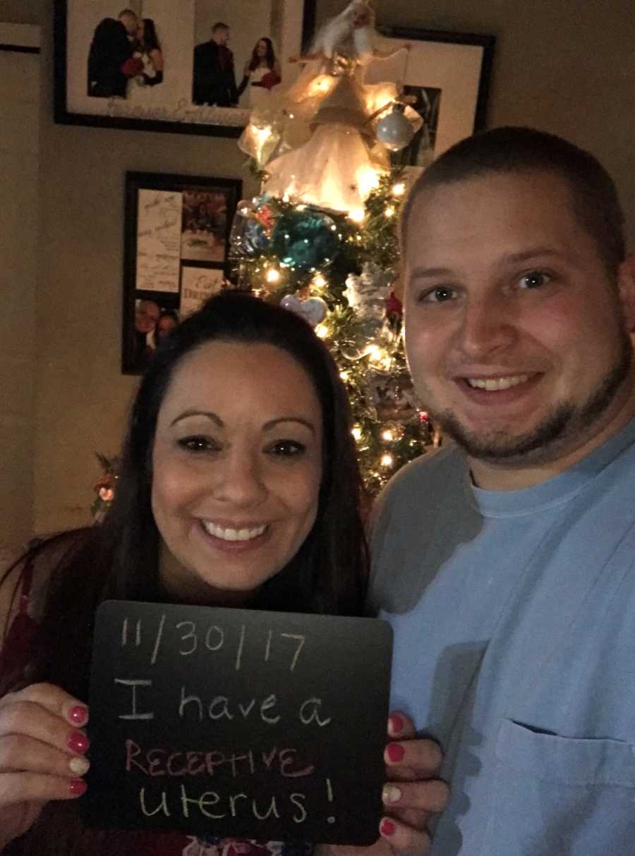 Wife who struggles with infertility holds up sign saying, "I have a receptive uterus" with husband in selfie