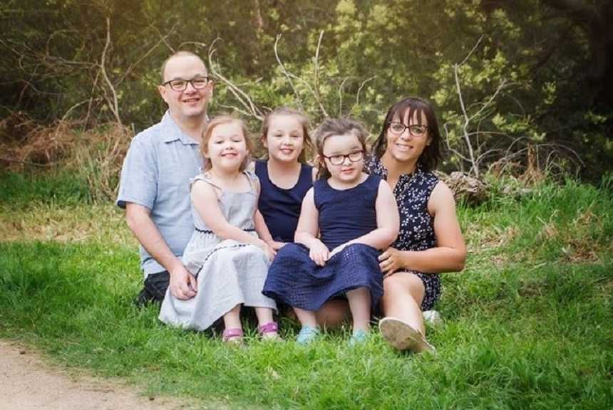 Little girl who had brain surgery smiles on moms lap next to her two sisters and dad as they sit on the grass