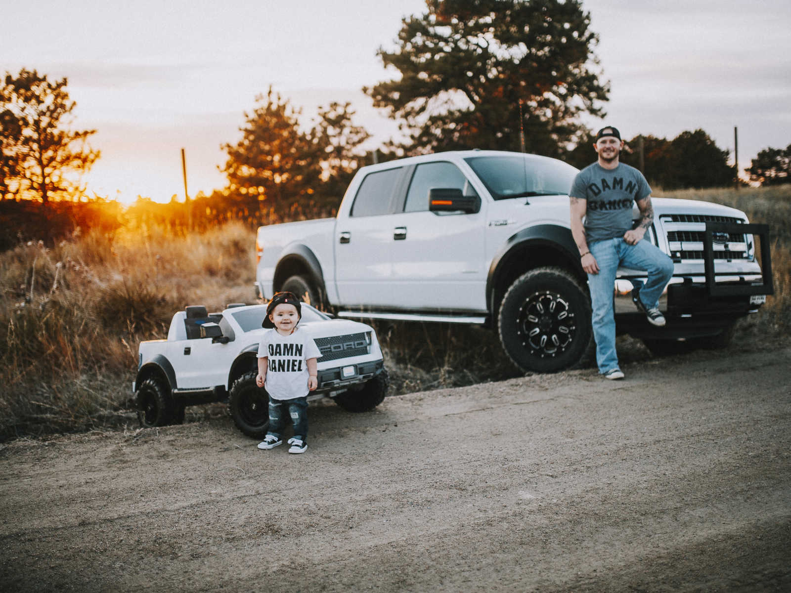 Father standing against his truck next to toddler son who is standing against his identical toy truck