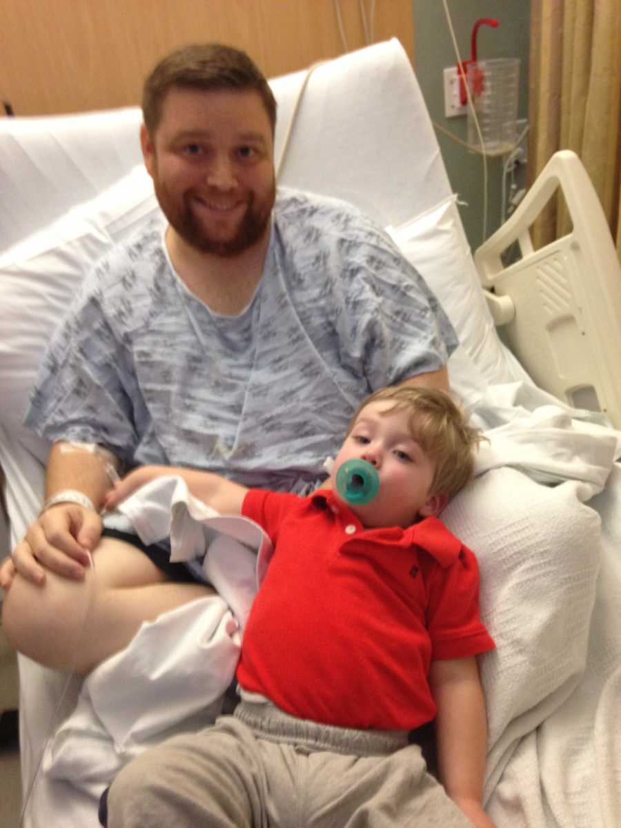 Man who needs kidney transplant sits up in hospital bed while toddler son leans against him