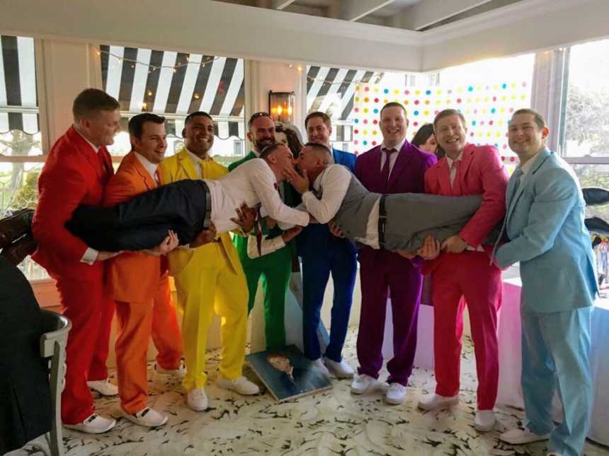 Men in rainbow colored suits hold grooms in the air while grooms kiss