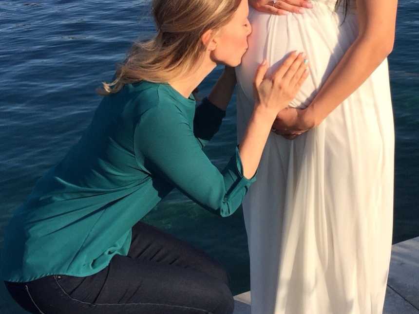 Woman crouches down on dock to kiss pregnant woman's stomach