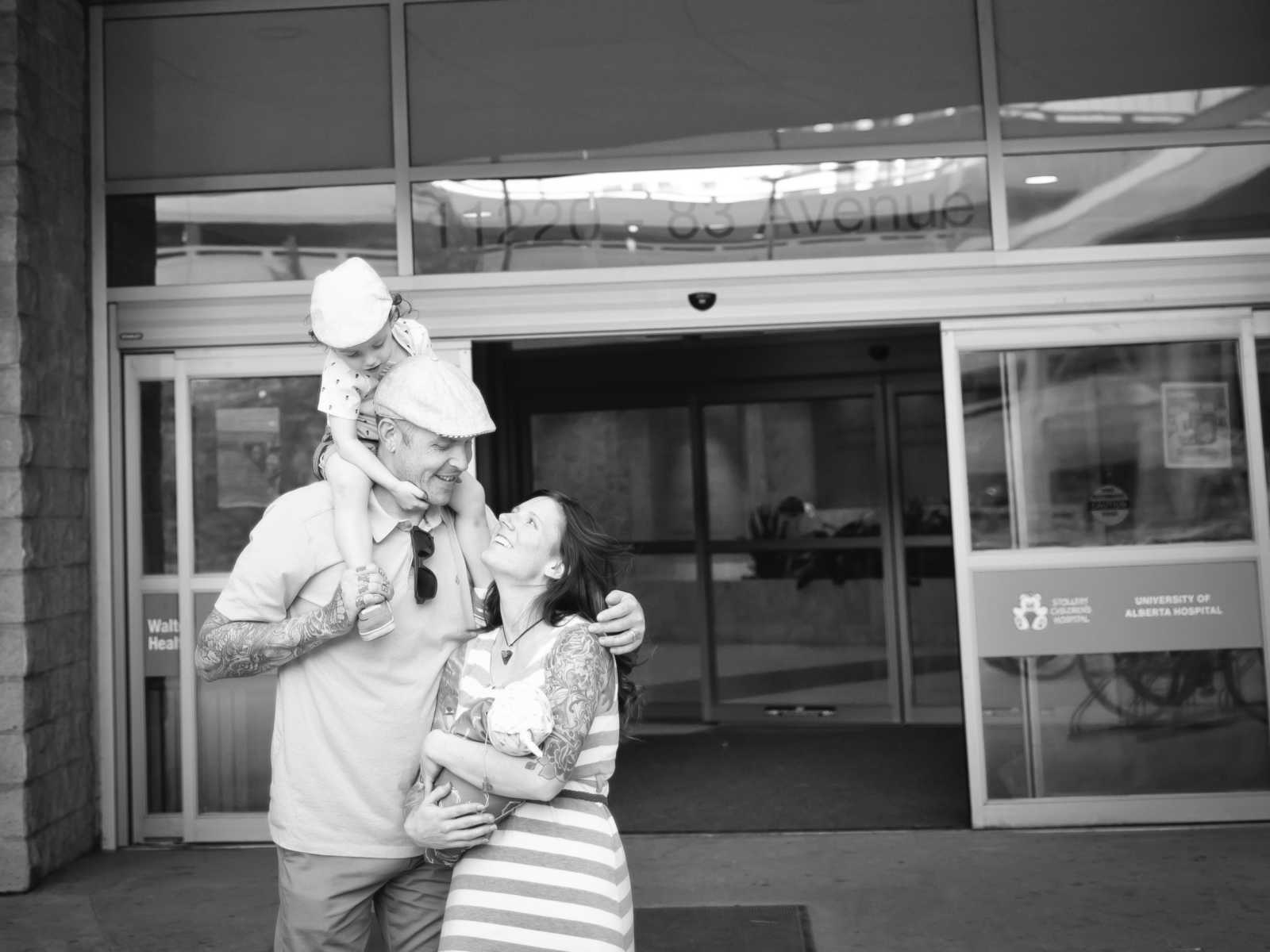 Mother holds newborn as she and her husband who has first born on his shoulders leave hospital