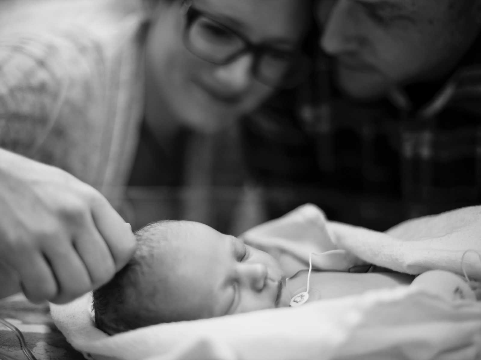 Father and mother stand over newborn sleeping while mother strokes newborn's head