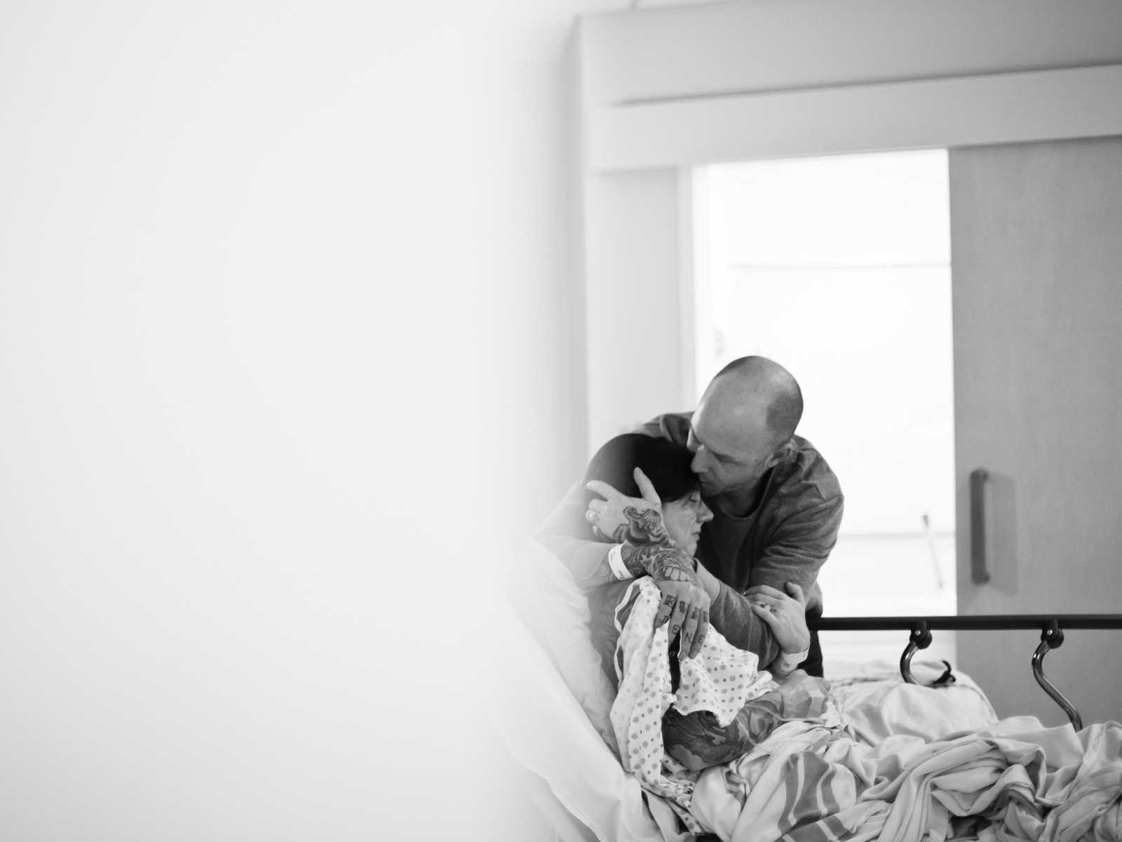 Woman who just gave birth sits up on hospital bed as husband kisses her fore head with his arms wrapped around her
