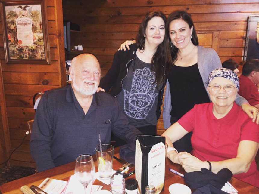 Woman with small cell cancer sits at restaurant table with husband and two granddaughters standing behind them