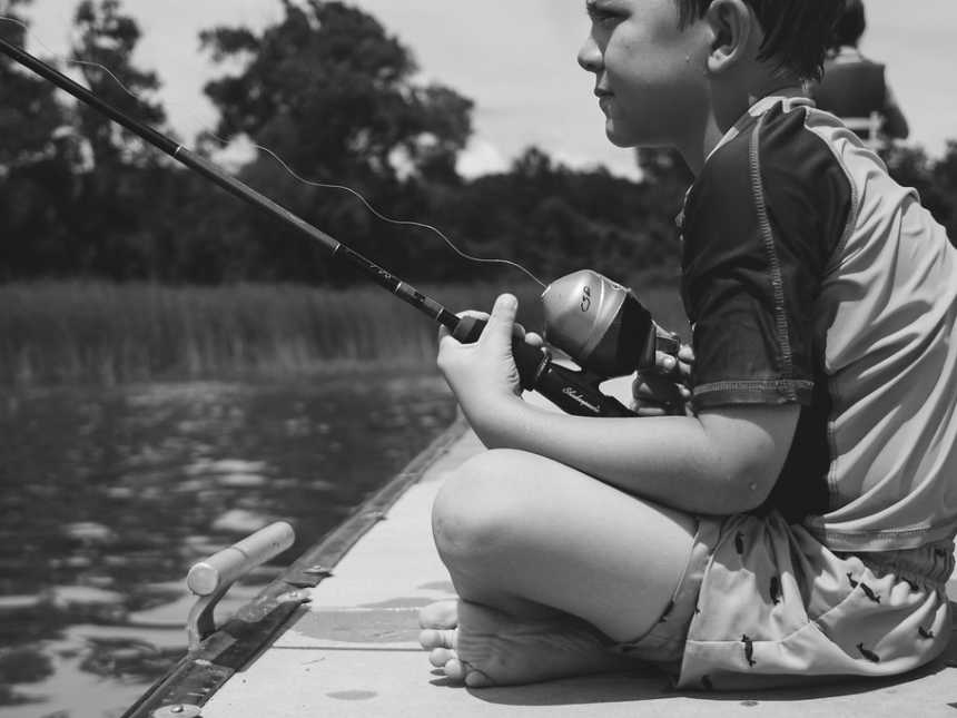 Little boy sitting on dock with fishing pole in hand looking out at body of water