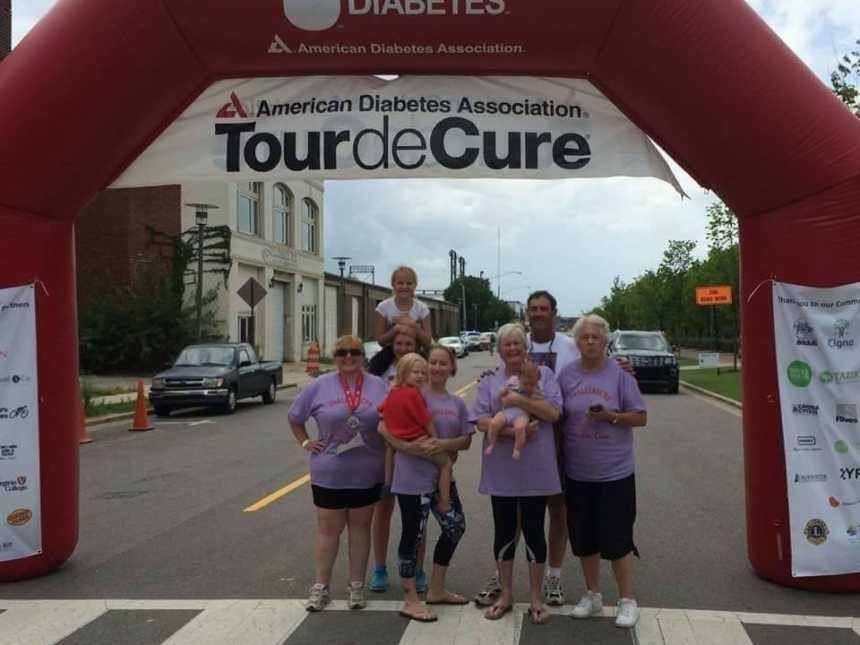 Wife and mother who has diabetes stands with family under pink finish like that says,"tour de cure"