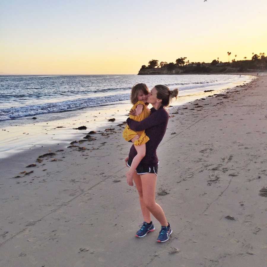 Mother holds daughter and kisses her on the cheek at shore of ocean