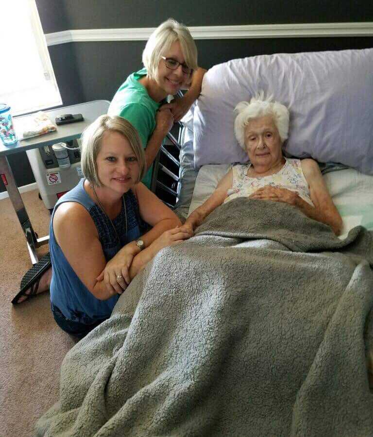 97 year old woman lays in hospital bed smiling with her granddaughters smiling at her side