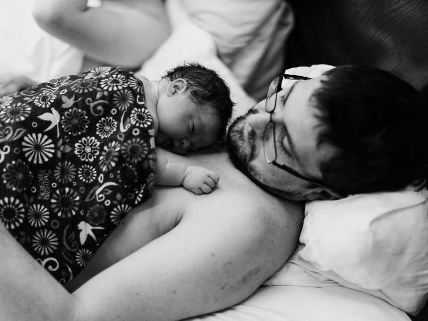 Father lays shirtless in bed with newborn on his chest