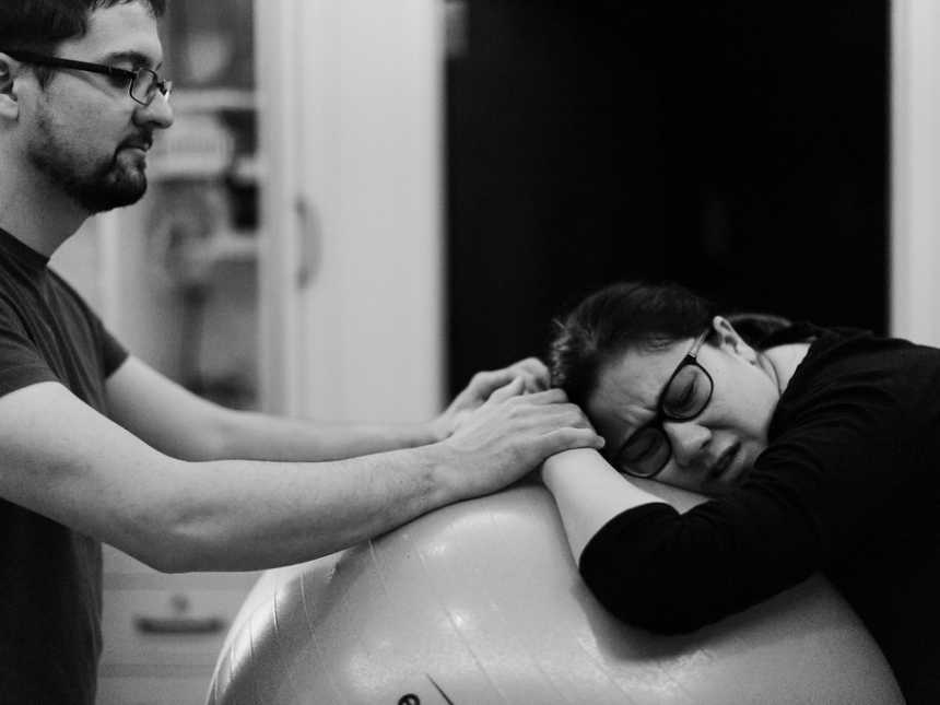 Pregnant woman leans head on medicine ball while husband holds her hands
