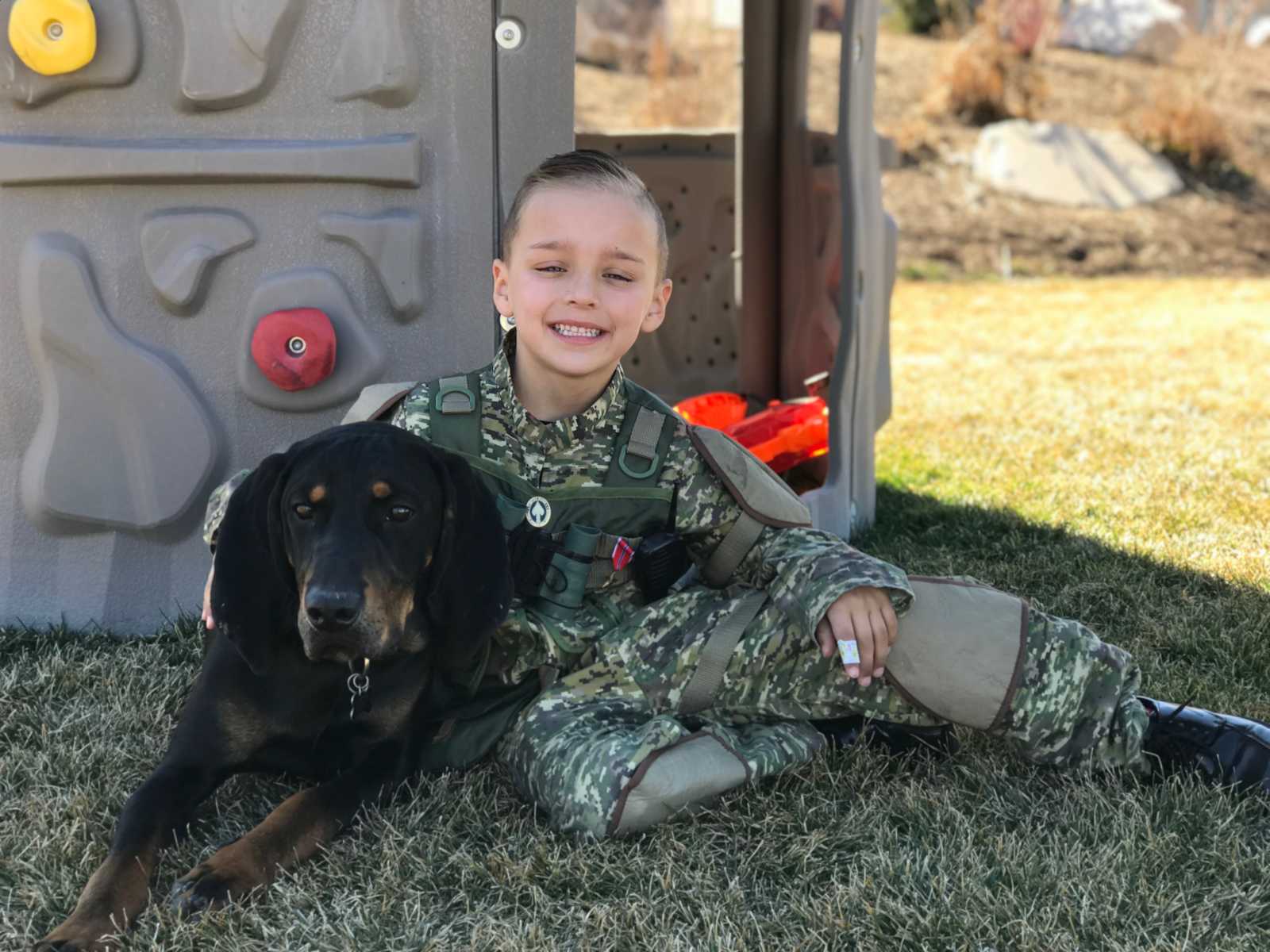 Little boy with leg tumors sitting in grass smiling with dog