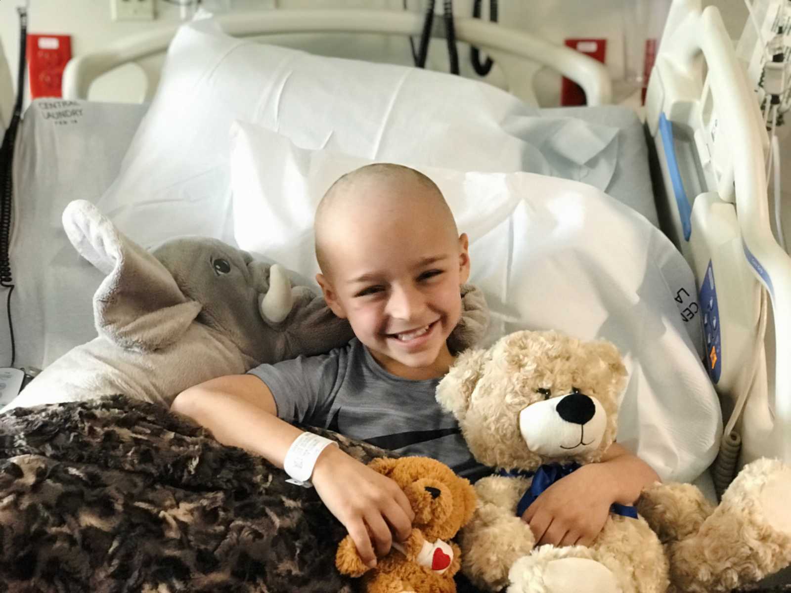 Boy who had leg tumors smiling in hospital bed with stuffed animals in his lap