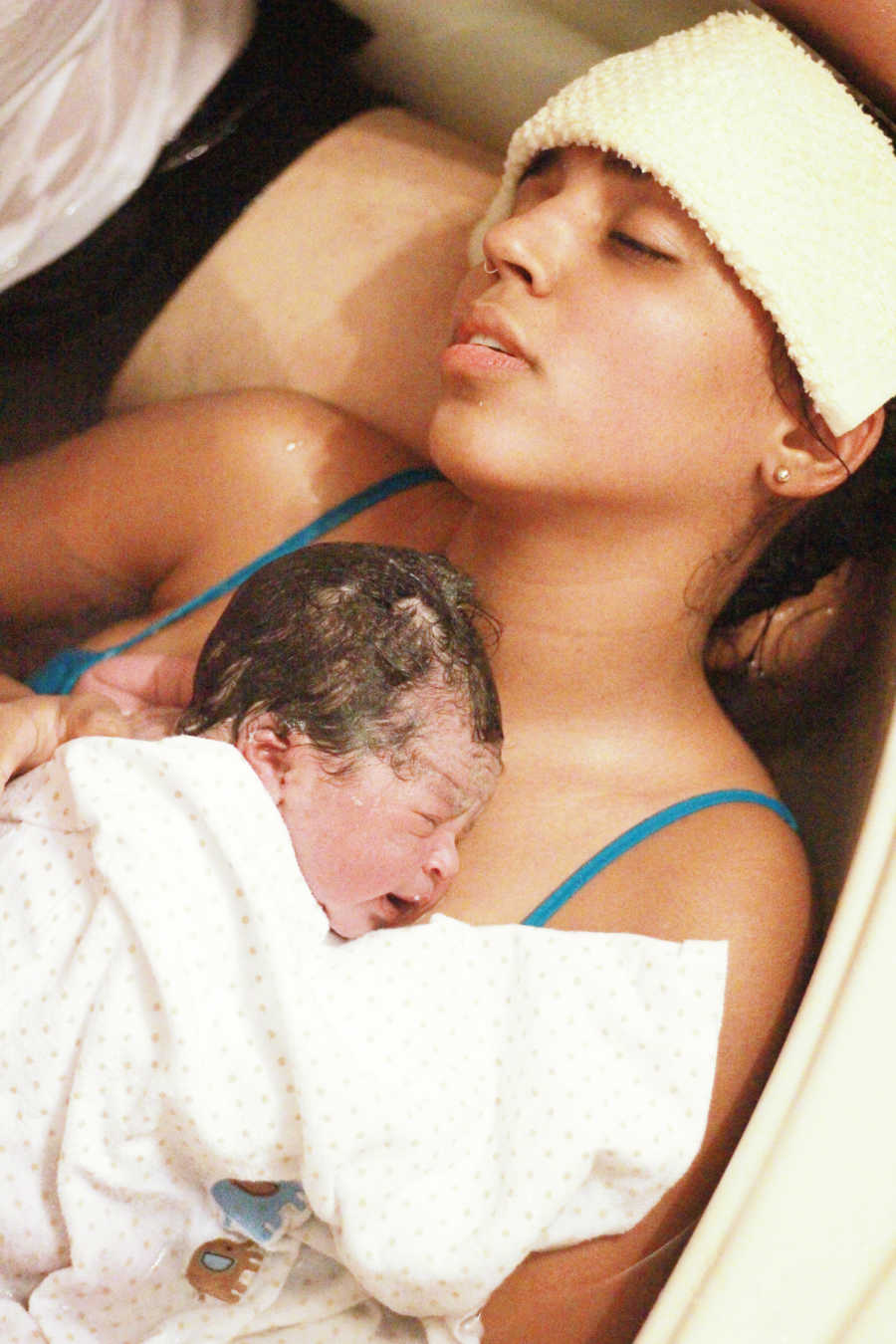 Woman who just had water birth lies with newborn swaddled in a towel to her chest with towel over her head