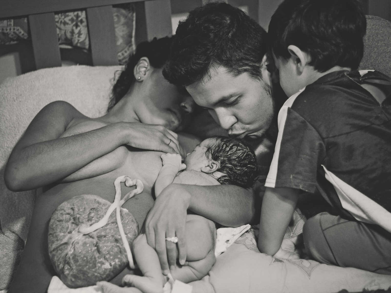 Shirtless mother who had water birth lies in bed breastfeeding while husband kisses newborn and son watches