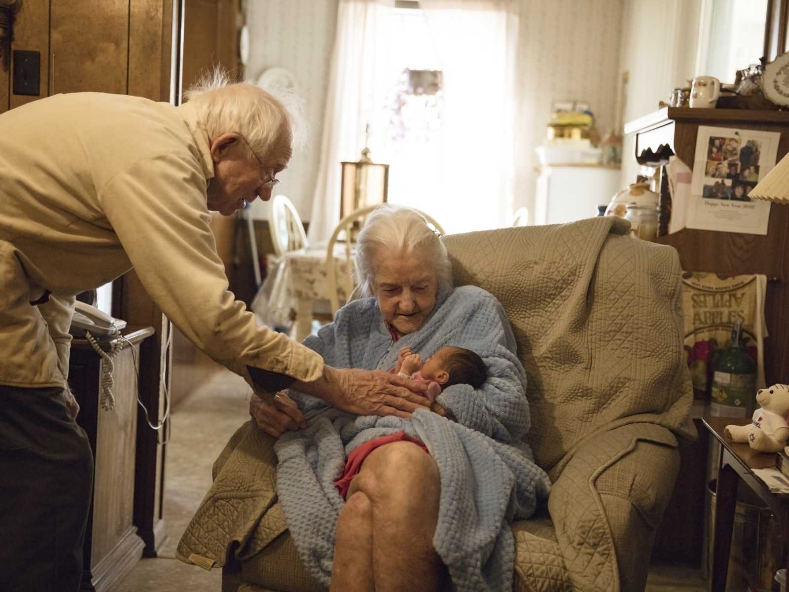 92 year old woman sits holding newborn who is her namesake in her lap while husband reaches over to touch newborn