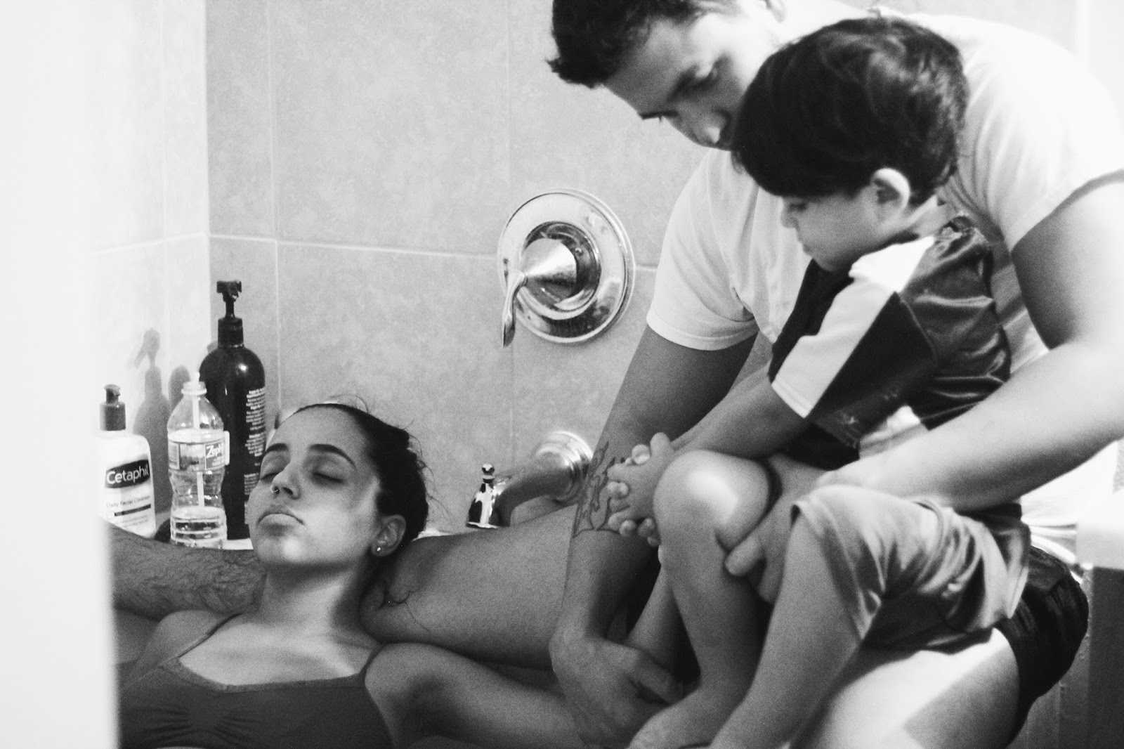 Woman giving water birth lies back in tub while husband sits on edge of tub with son in his lap
