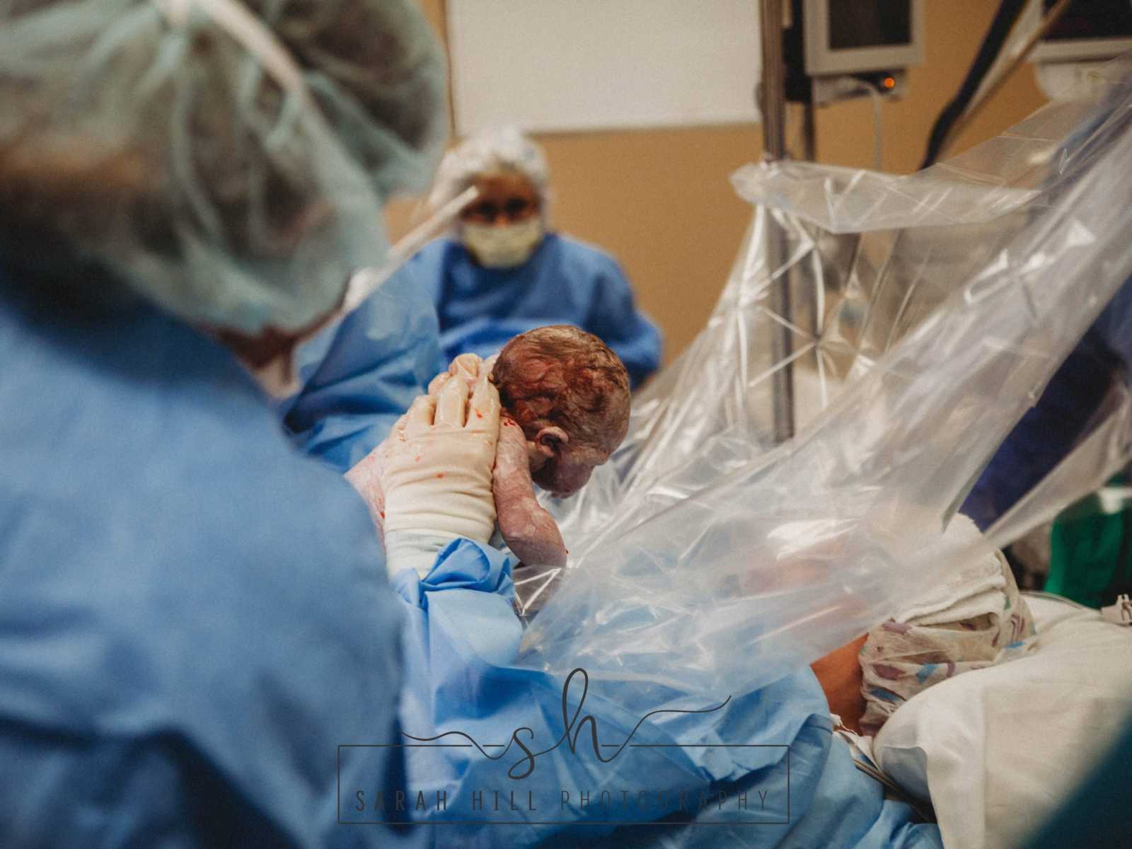 Woman who is also a midwife holds her newborn up at looks at it through surgical shield