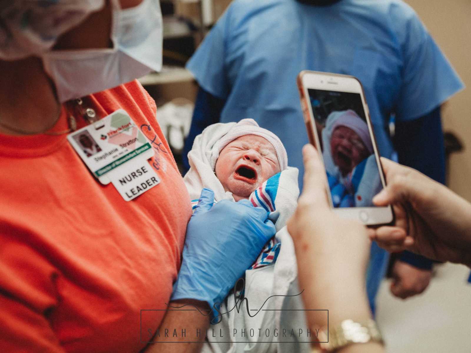 Nurse holding swaddled c-section newborn while someone takes picture on iphone