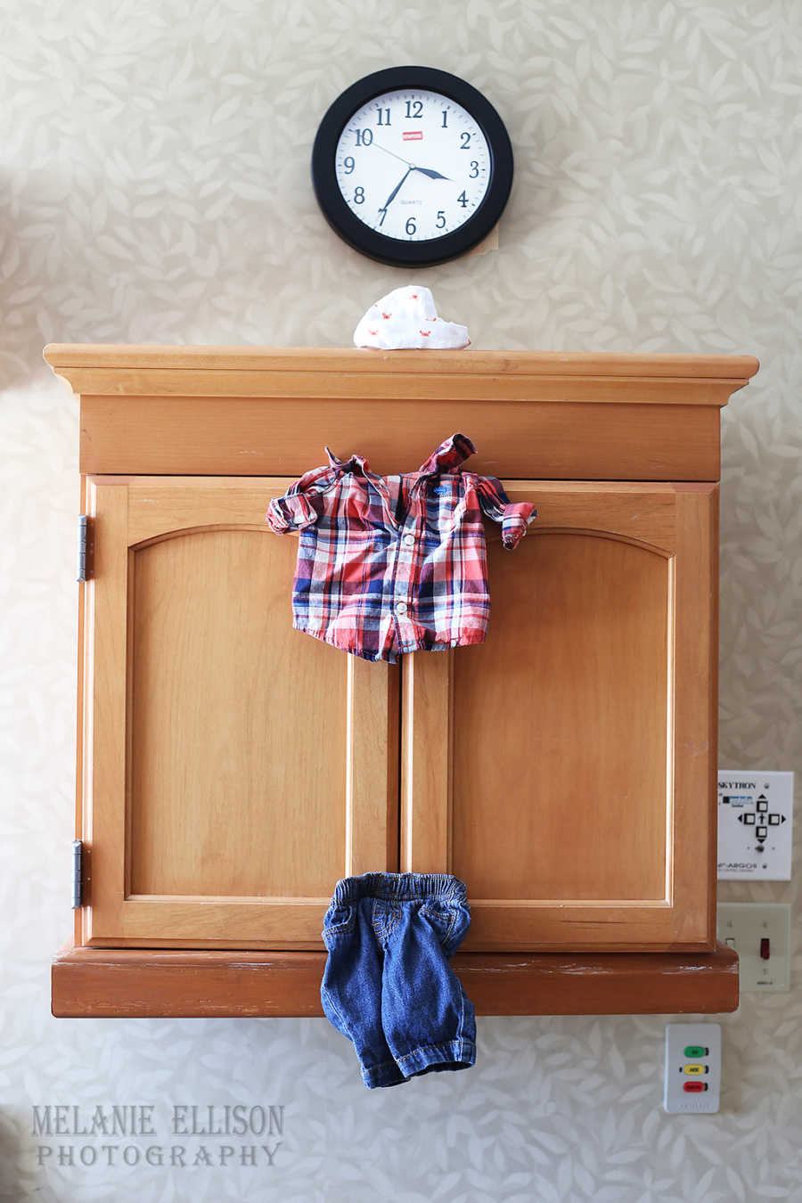 Wooden cabinet with baby boy shirt and pants stuck in doors with baby hat sitting on top of cabinet