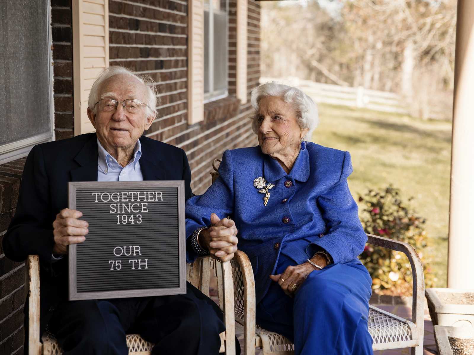 92 year old woman sits in chair holding hands with her husband who is holding sign that says, "together since 1943"