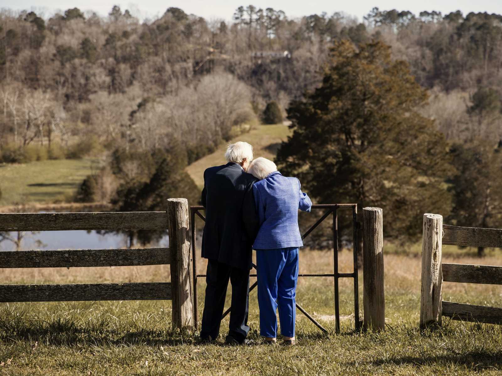 92-year-old woman leans her head on her husband's shoulders as they stand against iron gate looking out at grass field