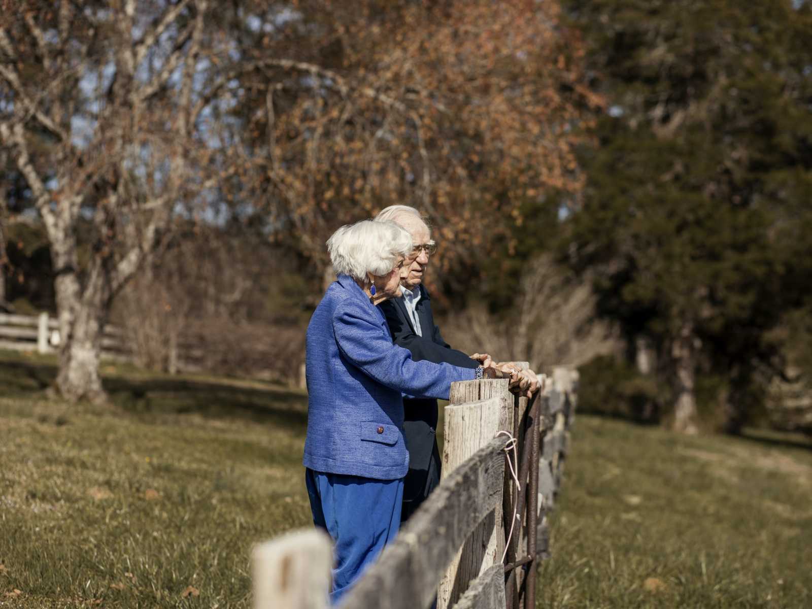 Husband stands with 92 year old wife leaning against wooden fence in their yard
