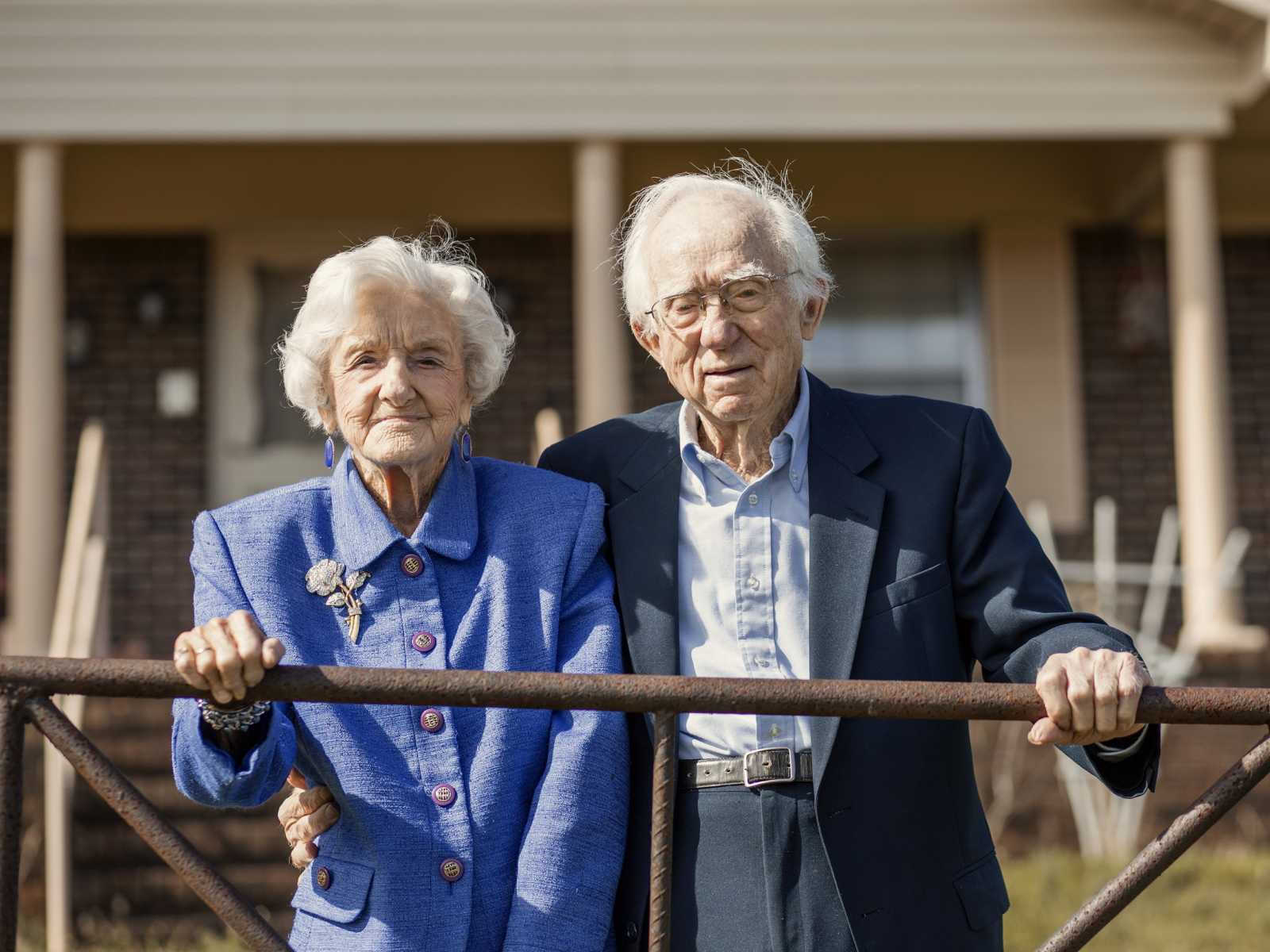 92 year old woman stands next to husband in formal attire in front of their home