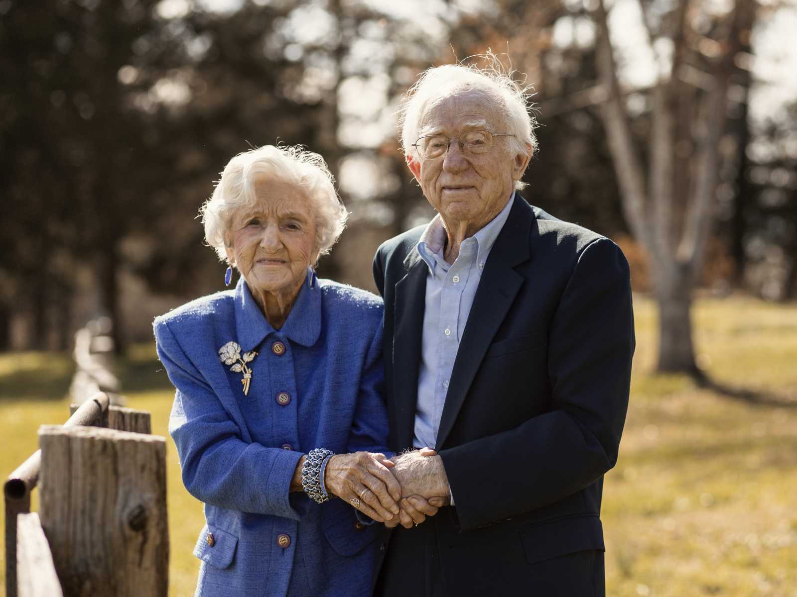 92 year old woman holds husbands hands as they smile in their yard next to wooden fence