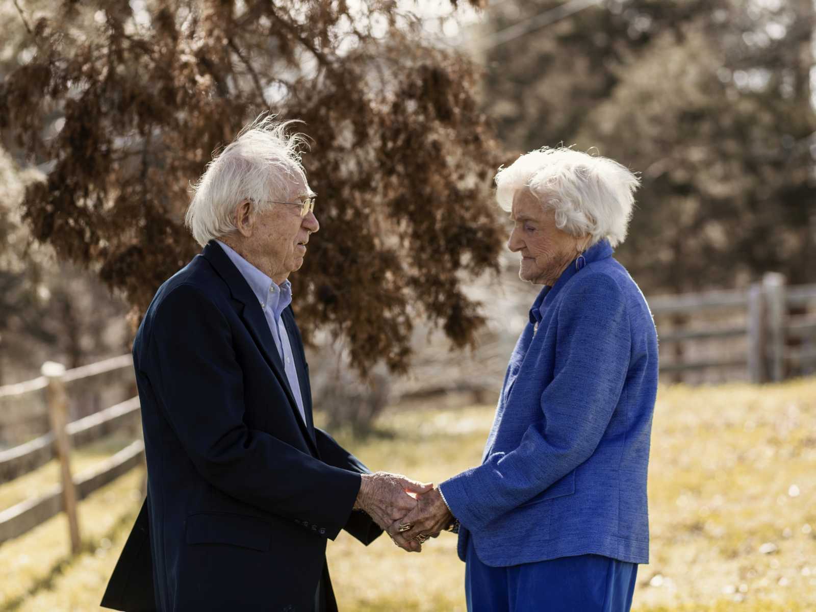 92 year old woman holds husbands hand as they stand face to face in their yard