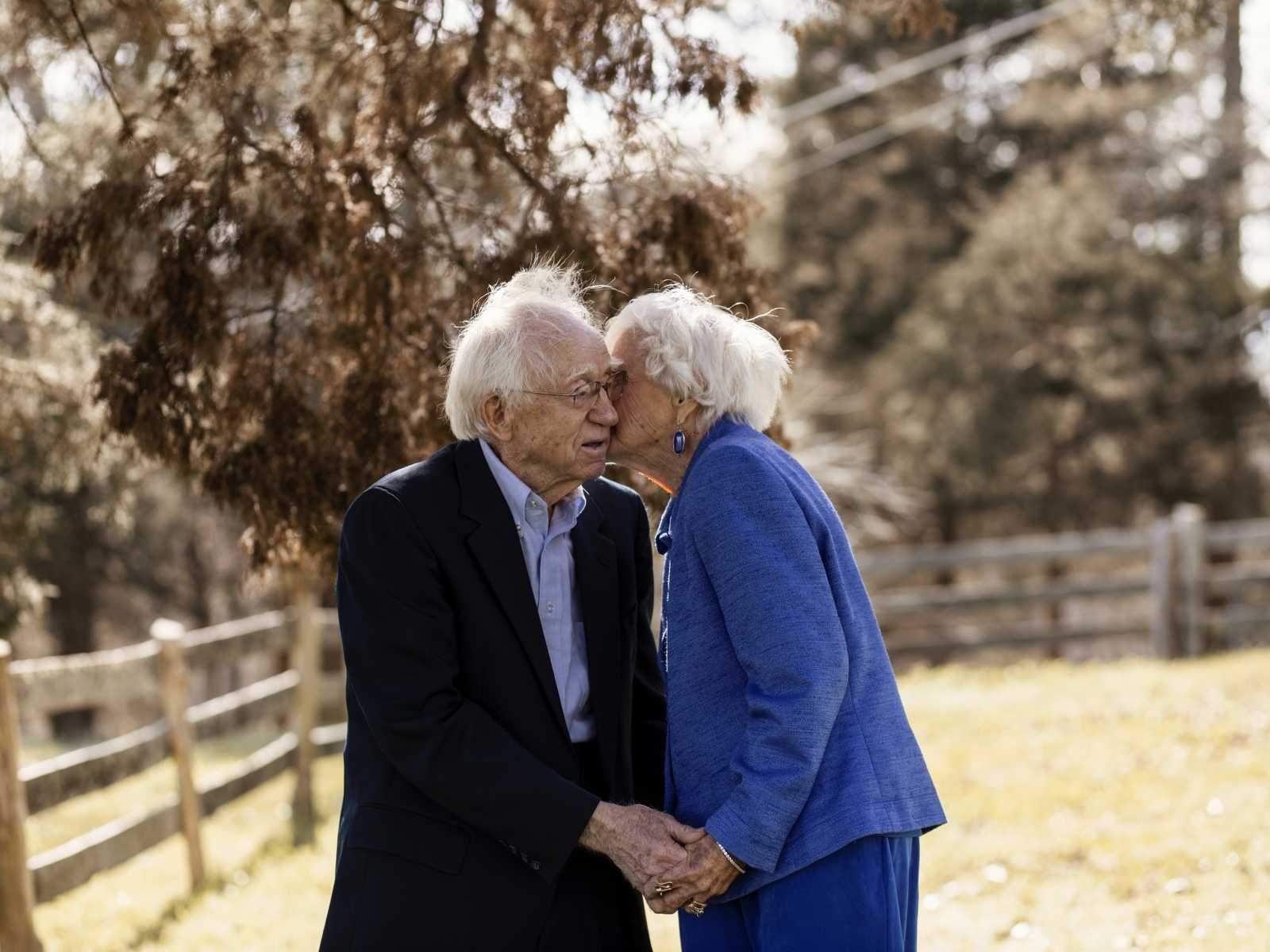 92 year old woman leans over to give her husband kiss on cheek as they hold hands