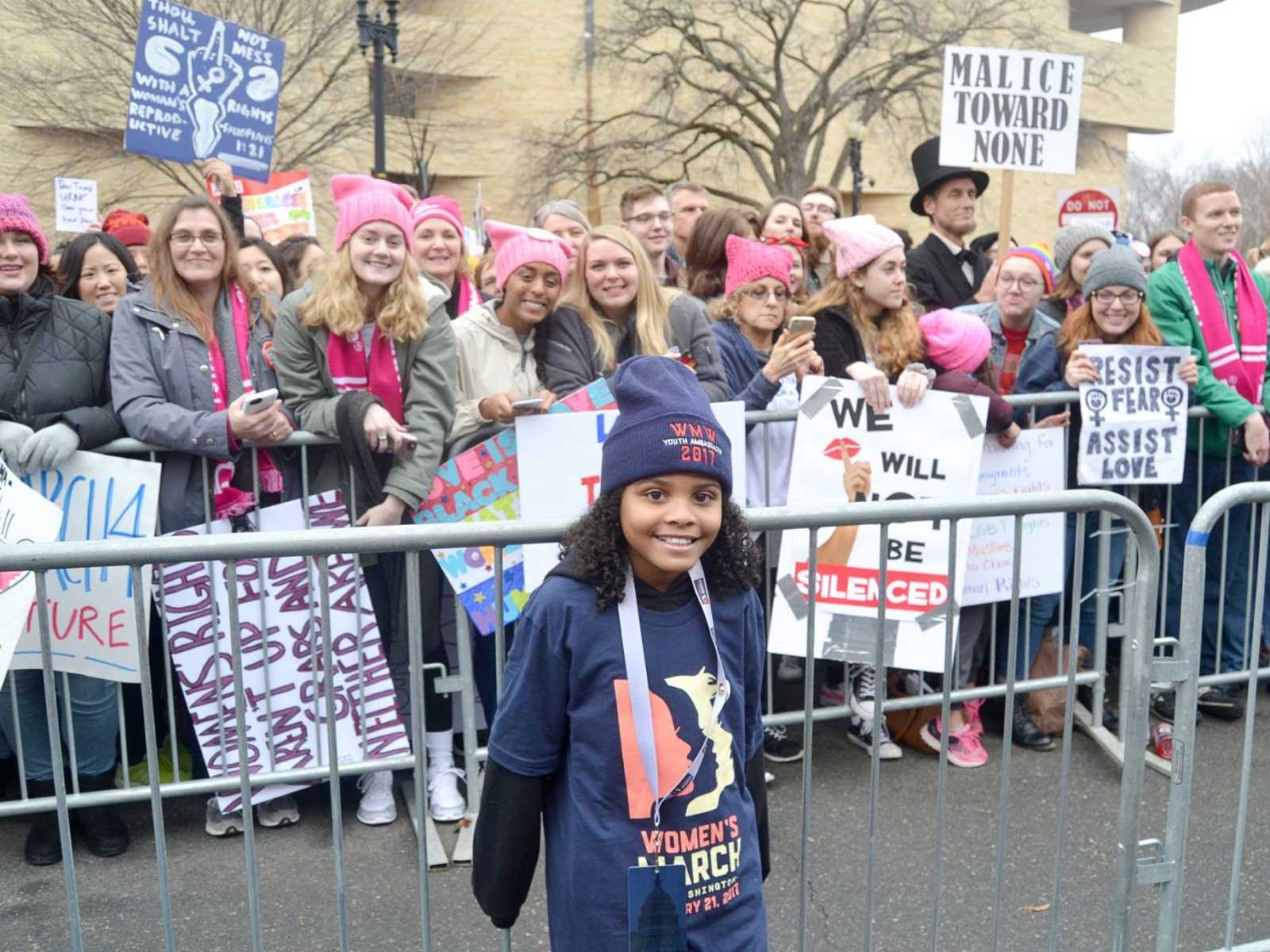 Little girl from Flint, Michigan smiles in front of fence in front of women holding posters at women's march