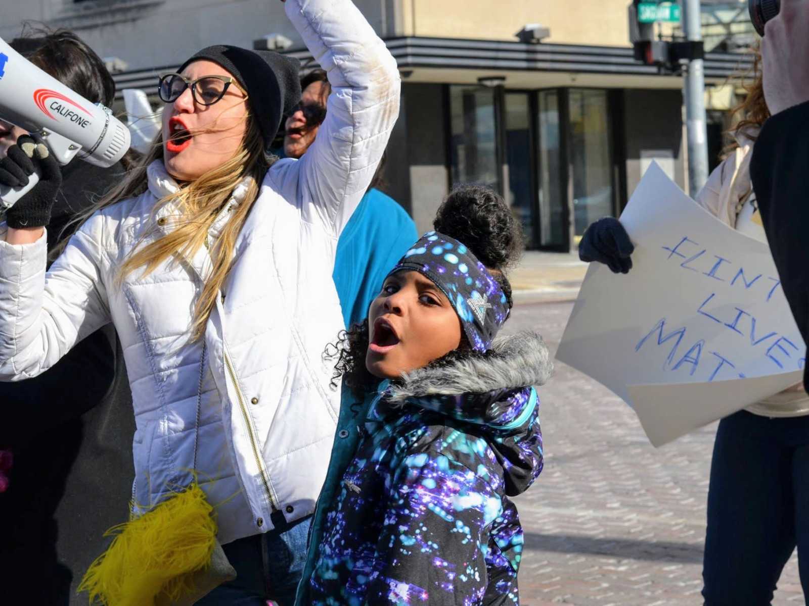Little girl and mother protesting the Flint water crisis and sign in background saying, "Flint Lives Matter"