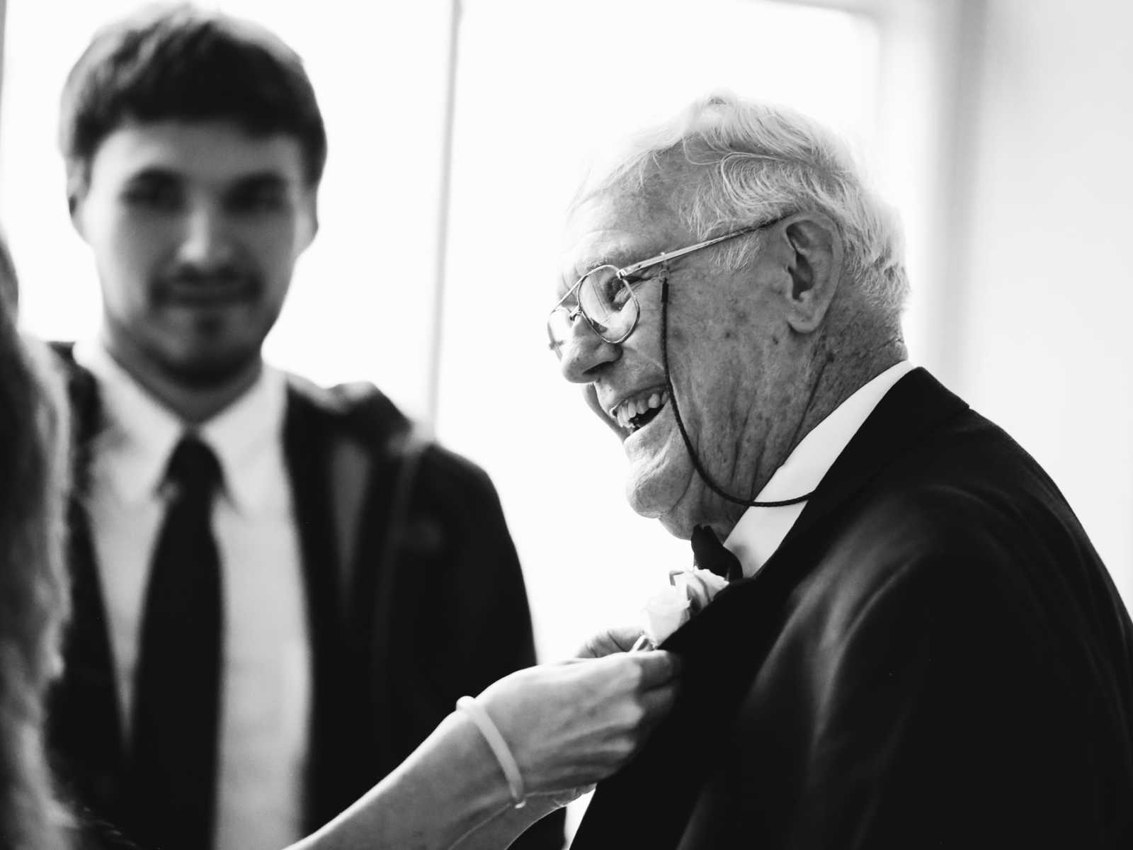 Elderly groom smiles as woman places boutonniere on him