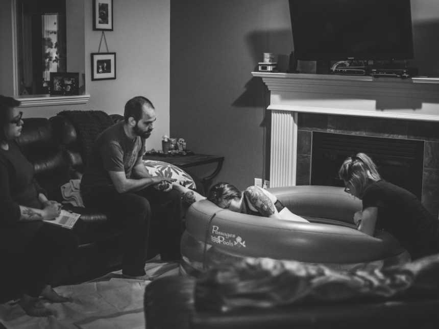 Pregnant woman sits in inflatable birthing pool with midwife, husband, and another woman on the side