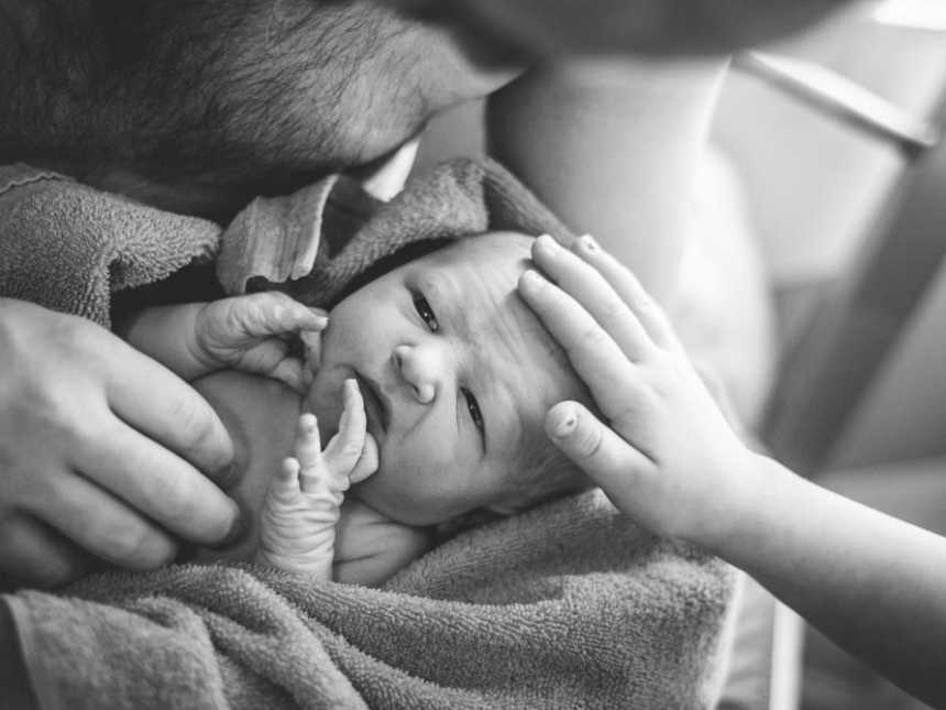 Shirtless father holding newborn in towel in his arms while firstborn touches baby's forehead
