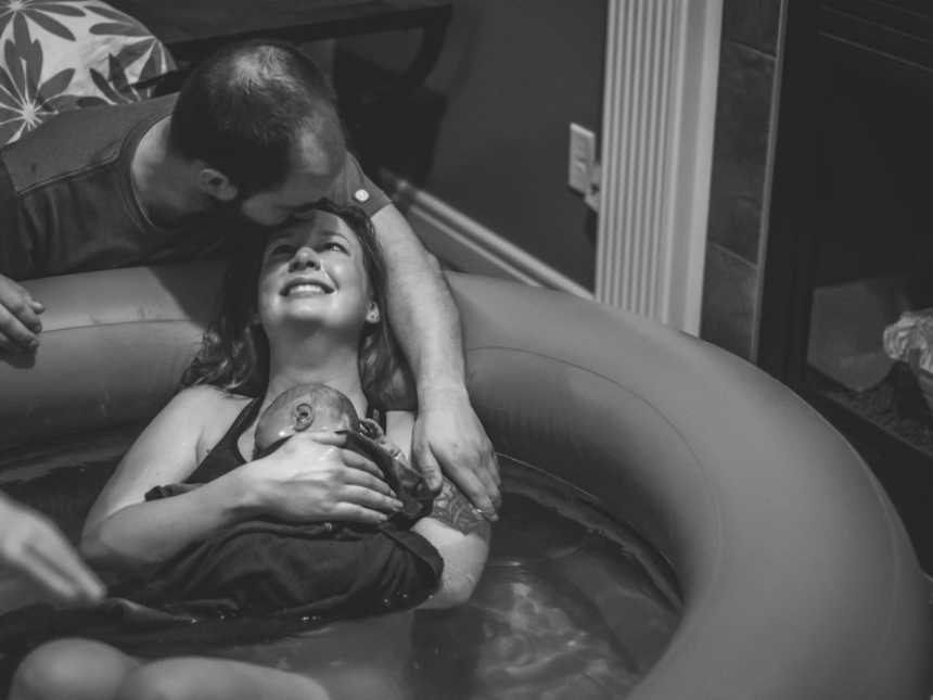 Woman smiles up at husband as she holds newborn in birthing pool as he kisses her fore head