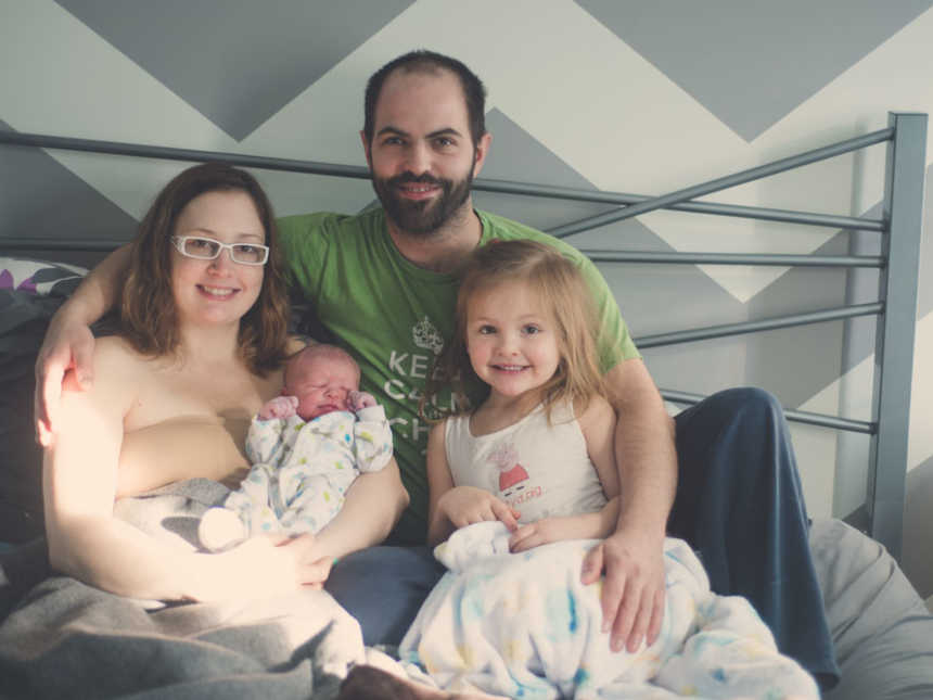 Mother holds newborn in arms in bed sitting next to husband and firstborn daughter