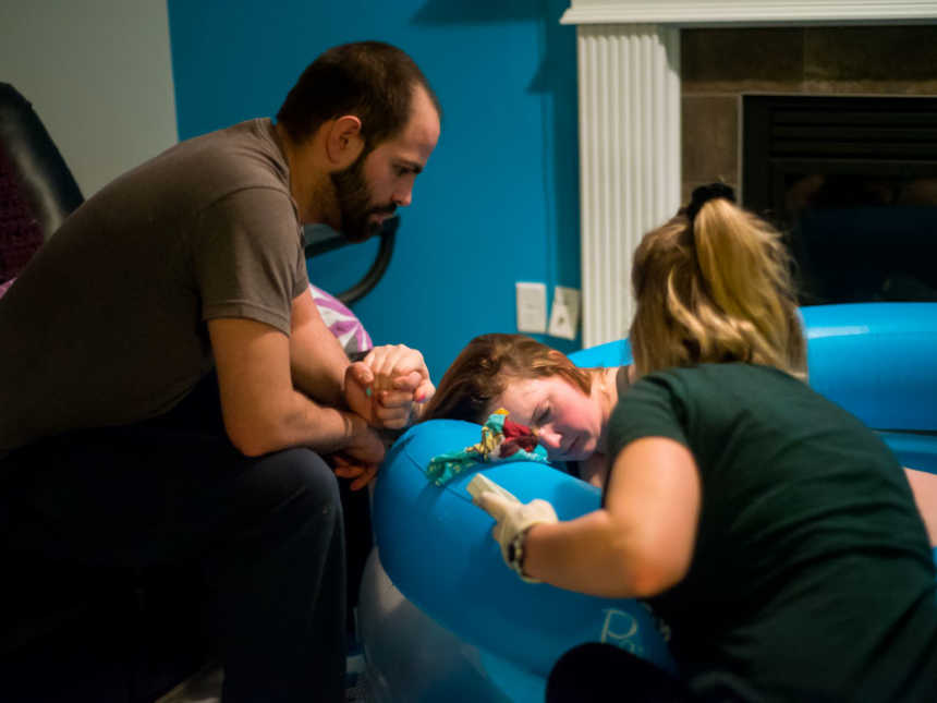 Pregnant woman leaning against birthing pool while holding husband's hand and midwife tends to her