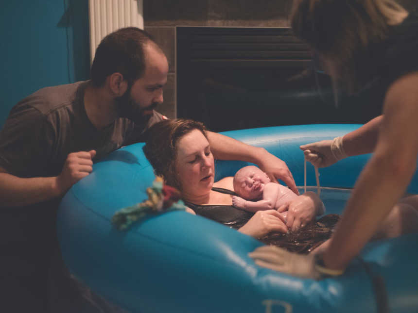 Woman leans against birthing pool while holding newborn who was encapsulated in sac while midwife holds umbilical chord