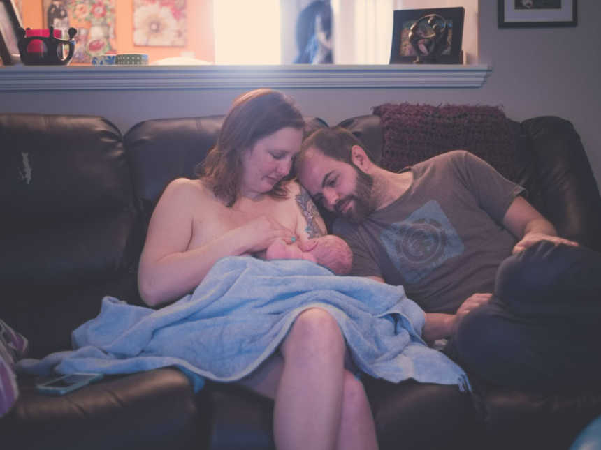 Woman breastfeeds newborn who was encapsulated in sac at birth on couch next to husband