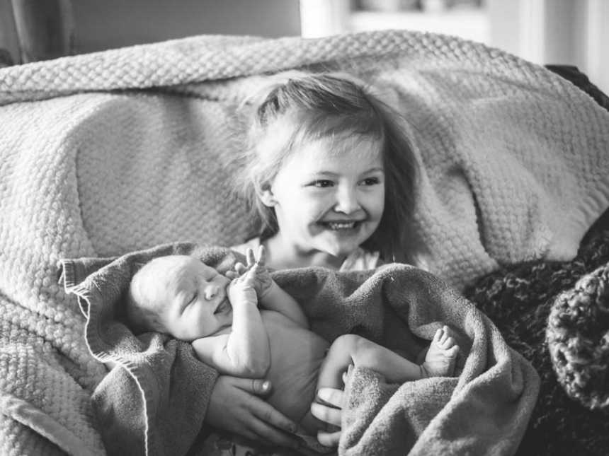 Firstborn smiles while sitting on couch holding baby brother who was encapsulated in sac at birth