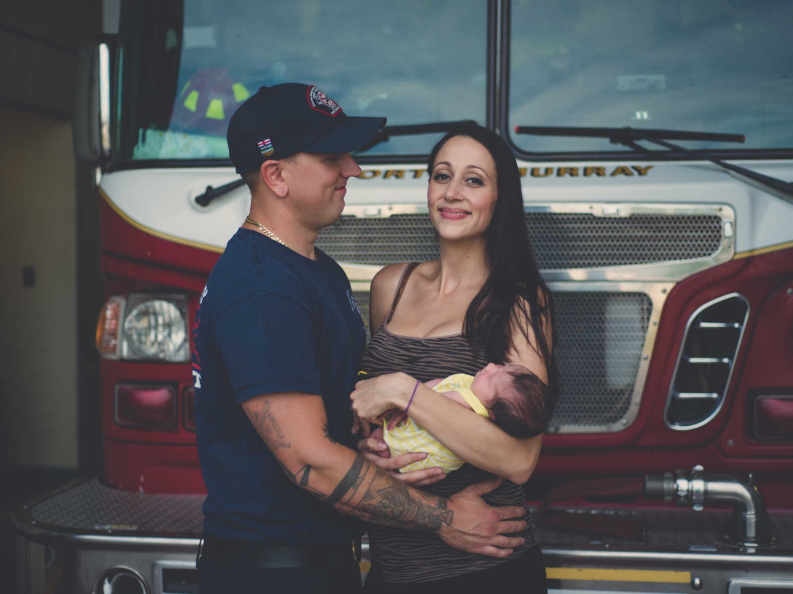 Mother and wife holds newborn in her arms next to fire fighter husband in front of fire truck