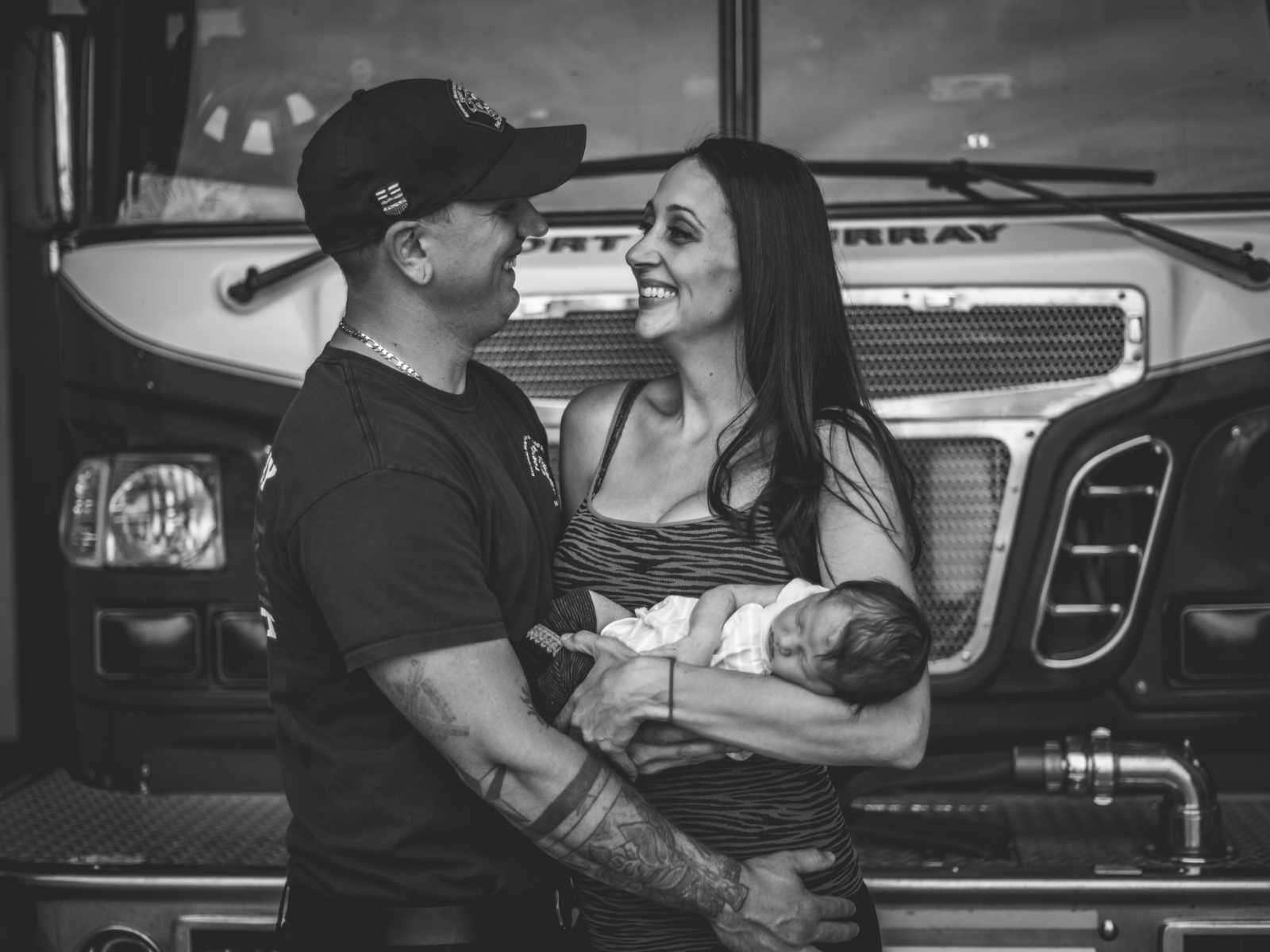 Wife smiles and looks in firefighter husband's eyes while holding newborn in front of fire truck