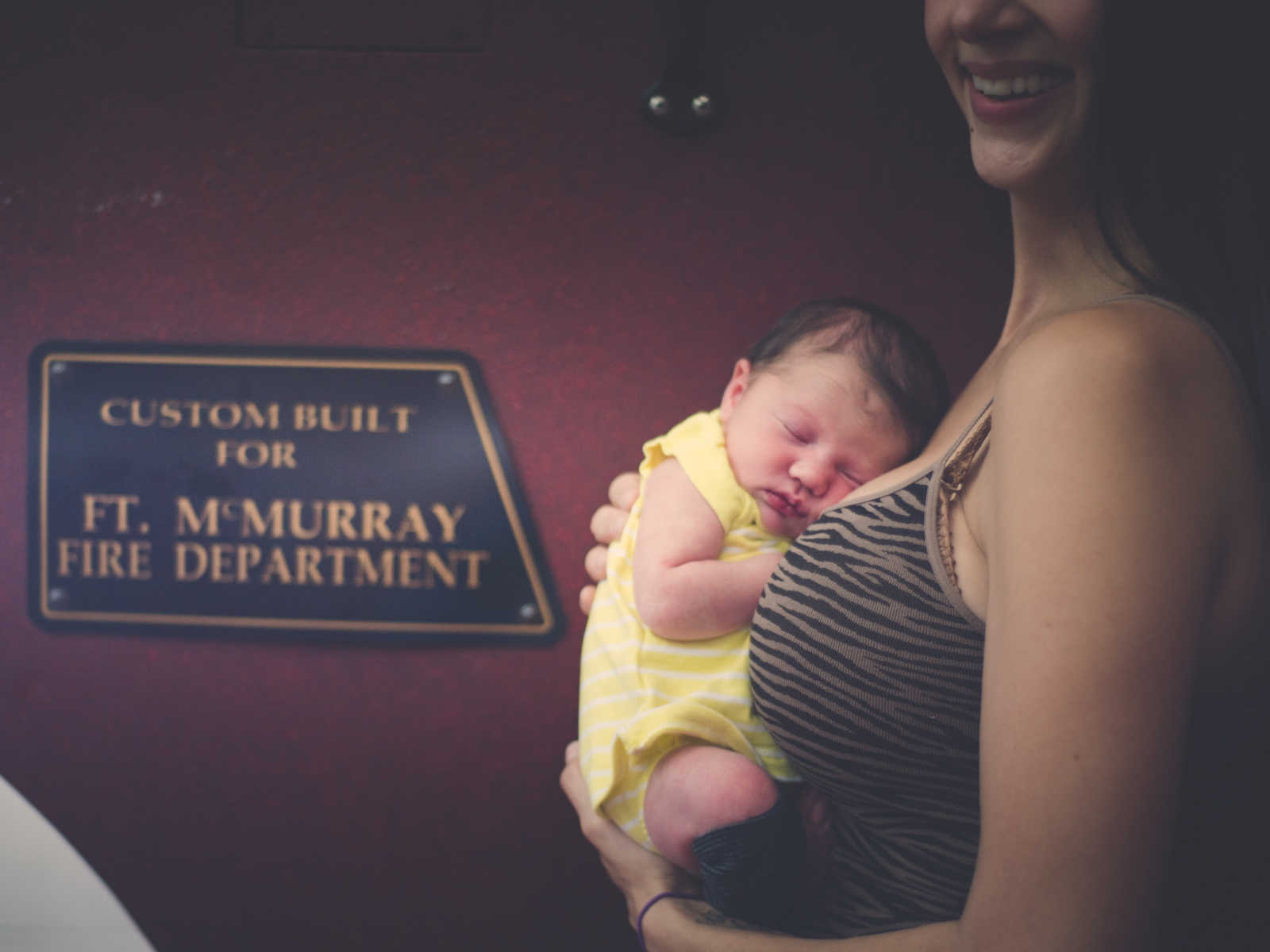 Close up of newborn of fire fighter being held in mother's arms next to sign saying, "Ft. McMurray Fire Department"