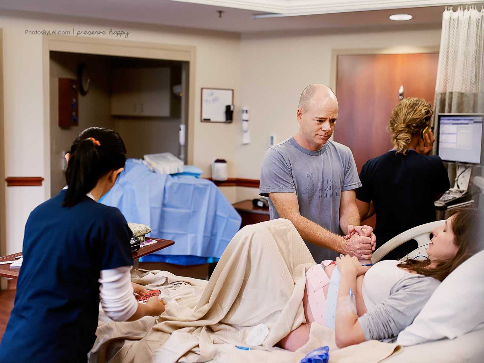 Pregnant woman lies in bed with eyes shut holding on to husbands hand while nurse tends to her