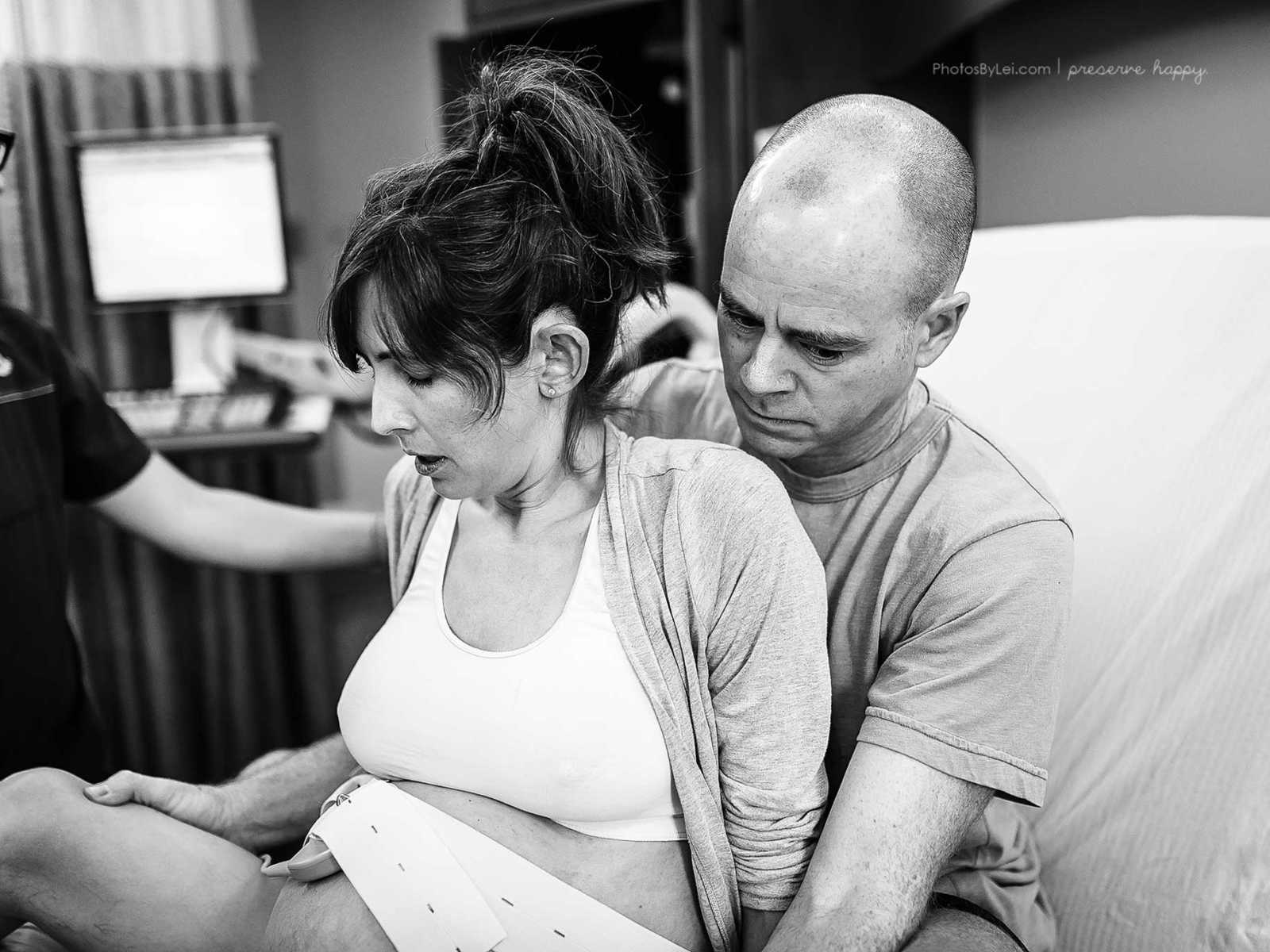 Husband sitting behind pregnant wife with fetal monitor on stomach in hospital bed for support