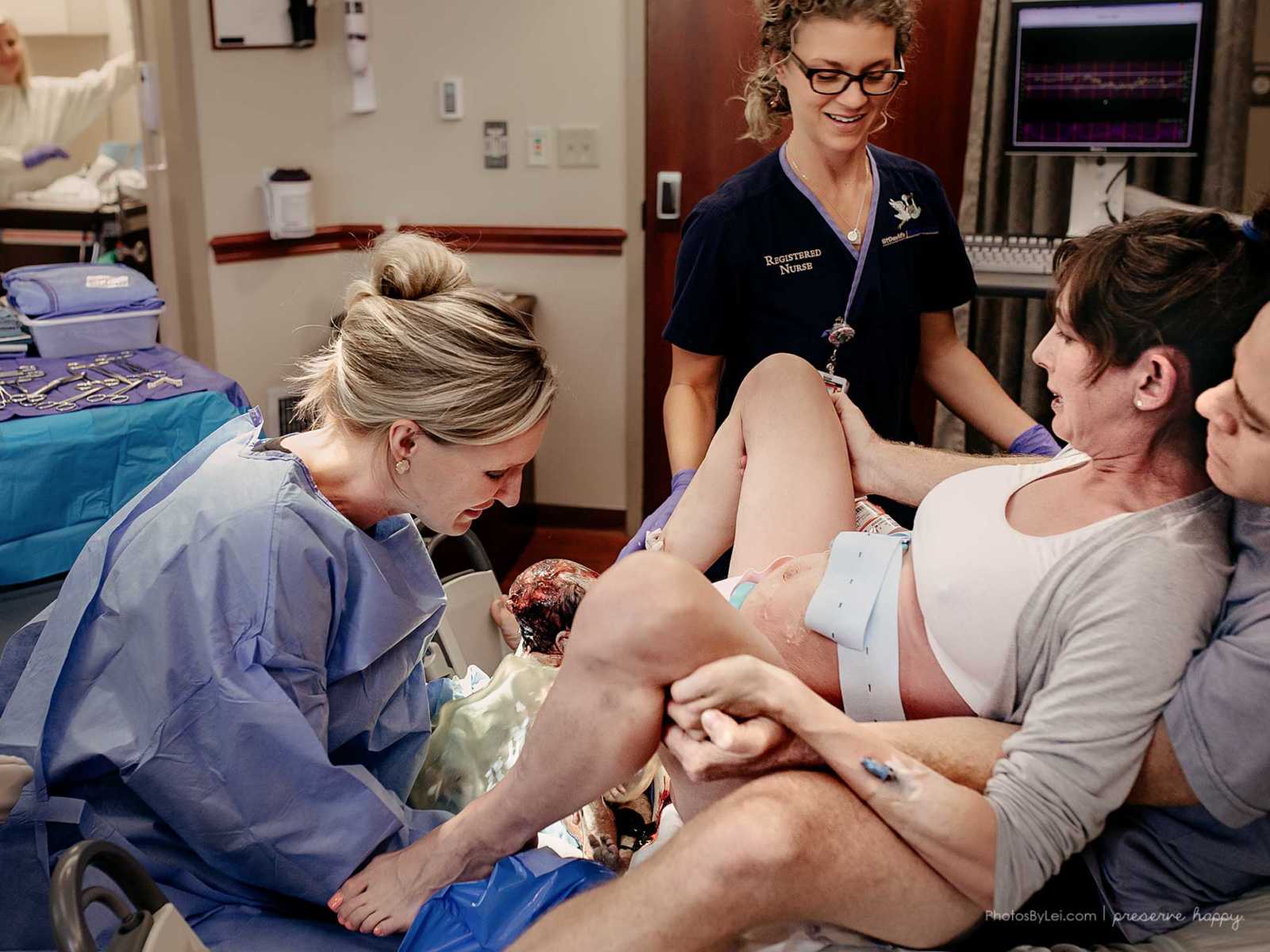 Pregnant woman giving birth with husband sitting behind her while OBGYN delivers baby and nurse holds woman's hand