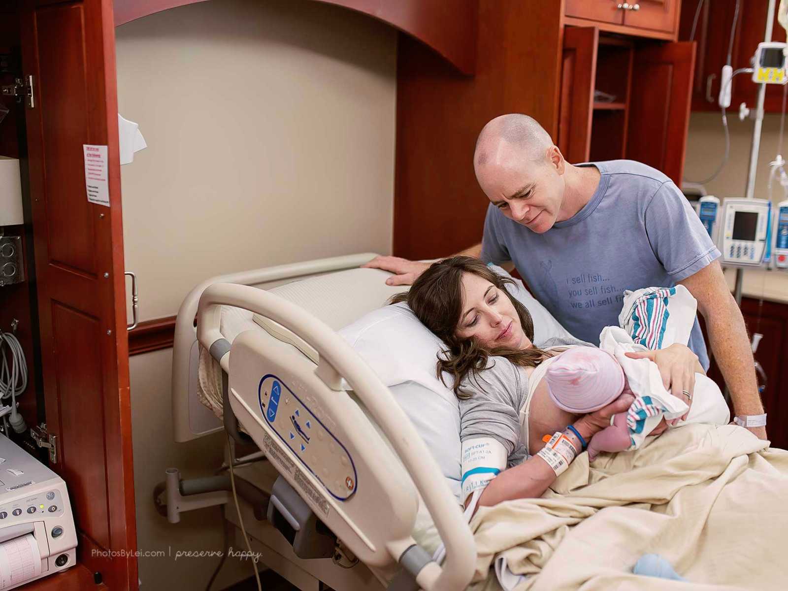 Father leans over hospital bed and looks at wife breastfeeding newborn who was covered in amniotic fluid